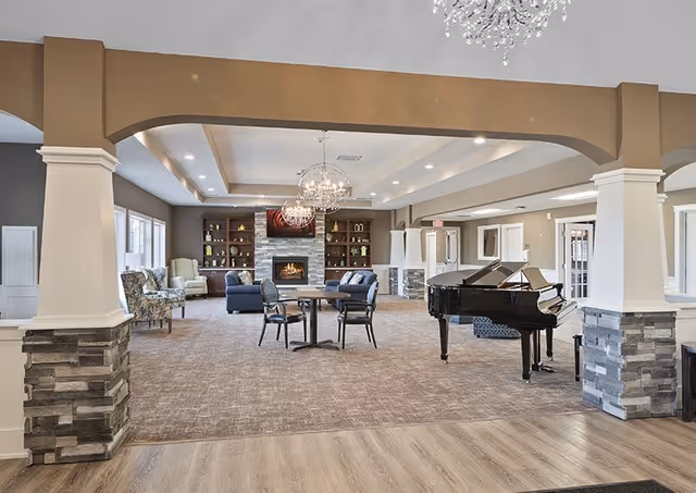 Spacious living room area with a grand piano on the right, a round table with chairs in the center, and a cozy seating area with sofas and armchairs near a stone fireplace. The room features chandeliers, large windows, and decorative columns with stone bases.