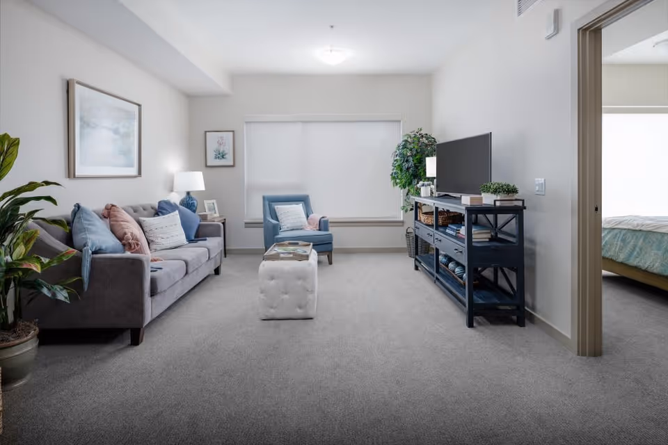 A bright living room with a gray sofa adorned with blue and pink pillows, a blue armchair, a white tufted ottoman, a TV on a dark blue console table, and a large window with a white blind. There are framed pictures on the walls, a potted plant on the left, and a doorway leading to a bedroom with a bed covered in a light blue patterned bedspread.