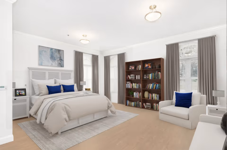 A bright and spacious bedroom featuring a large bed with beige bedding and blue accent pillows, two bedside tables with lamps, a wooden bookshelf filled with books and framed photos, two armchairs with blue pillows, large windows with gray curtains, and light wood flooring.