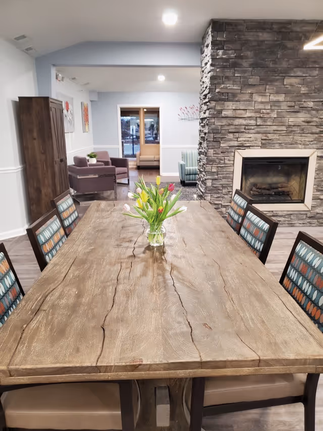 A long wooden dining table with a vase of colorful tulips in the center, surrounded by chairs with patterned upholstery. In the background, there is a stone fireplace on the right and a seating area with armchairs and a sofa. The room has light-colored walls and recessed ceiling lights.