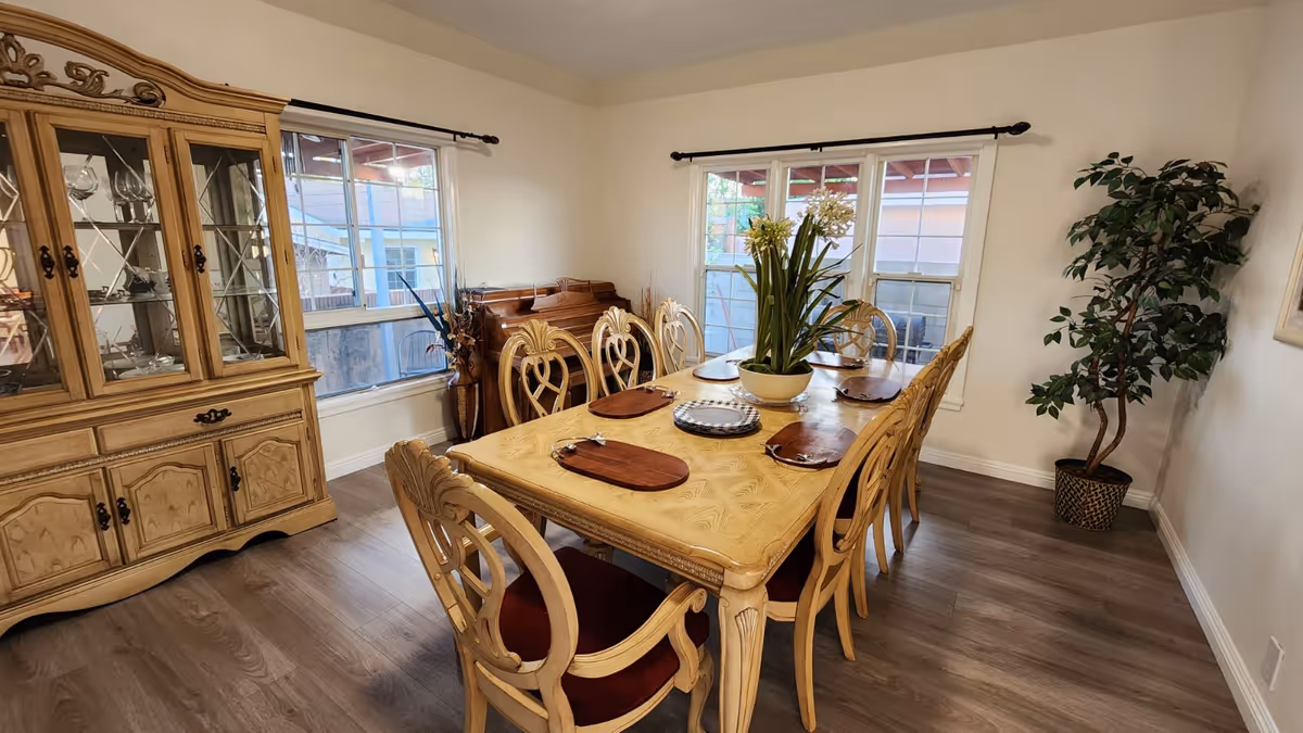 A dining room with a wooden dining table set for six, featuring a centerpiece with a potted plant. The room has wooden flooring, two windows with black curtain rods, a wooden china cabinet displaying glassware, a piano in the corner, and a tall potted plant near the wall.