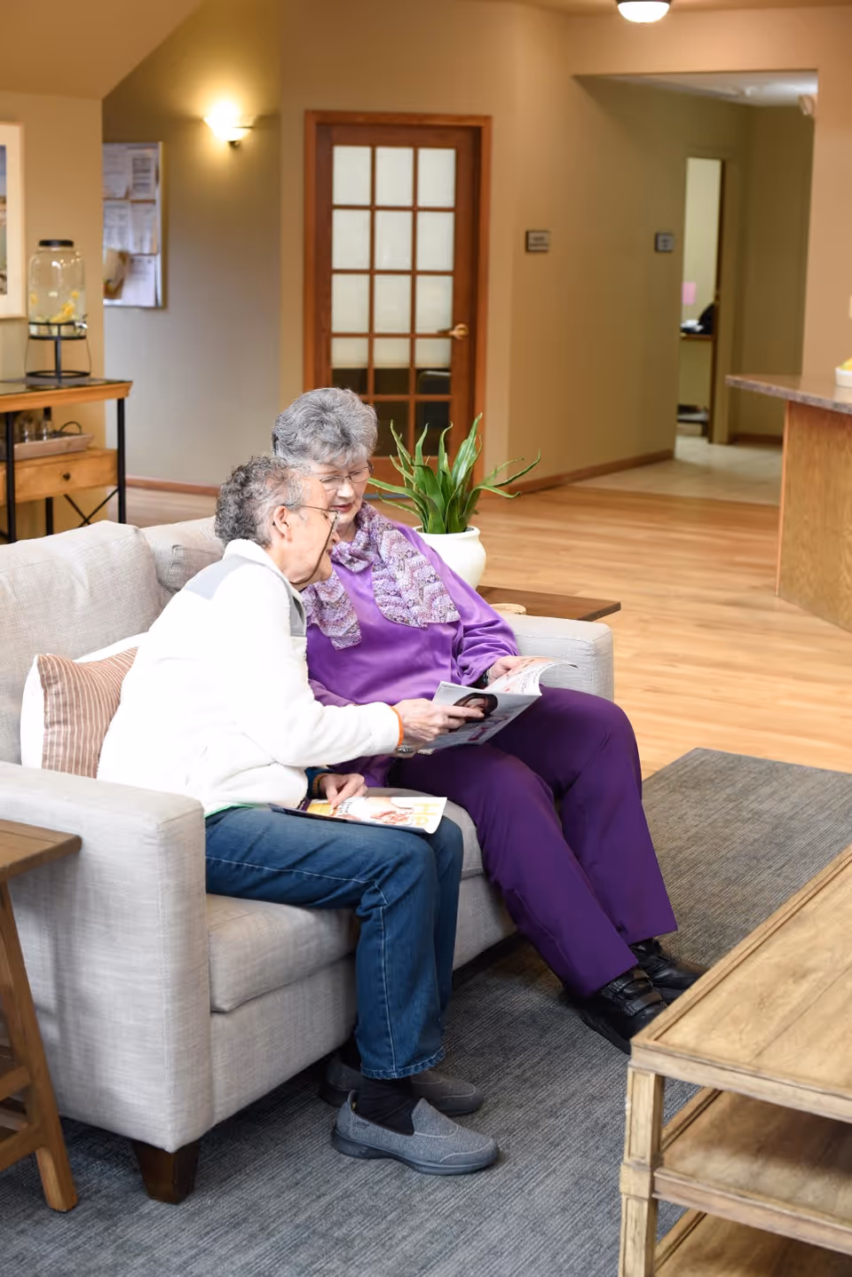 Two elderly women sitting on a light gray couch in a well-lit living area, looking at a magazine together. The room has wooden floors, a wooden coffee table, a potted plant, and a door with glass panels in the background.