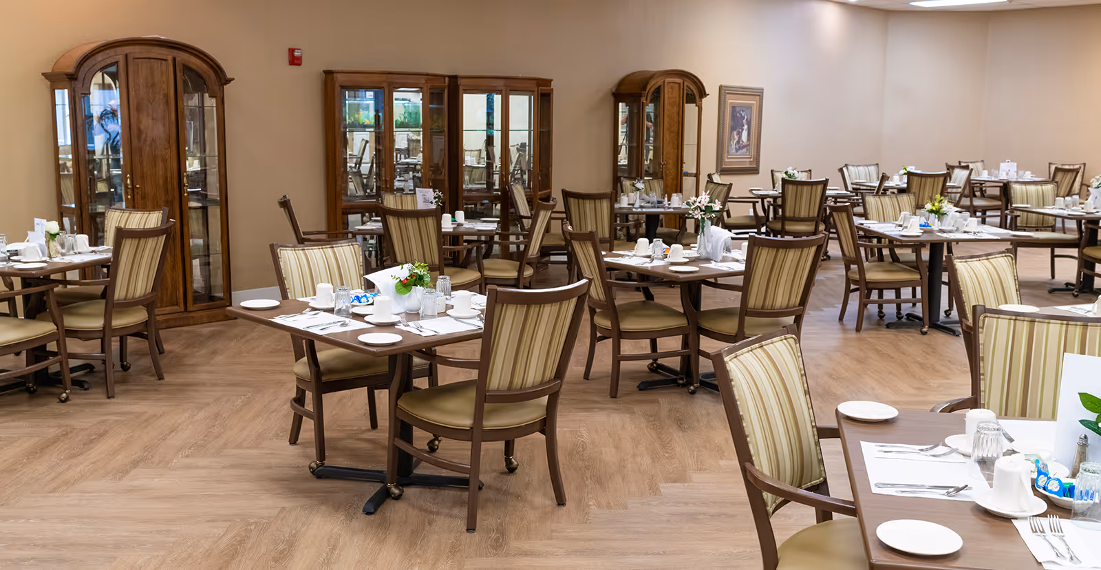 A spacious dining room with multiple tables set for meals, each table surrounded by wooden chairs with striped cushions. The room features wooden display cabinets along the walls and a light wood floor, creating a warm and inviting atmosphere.
