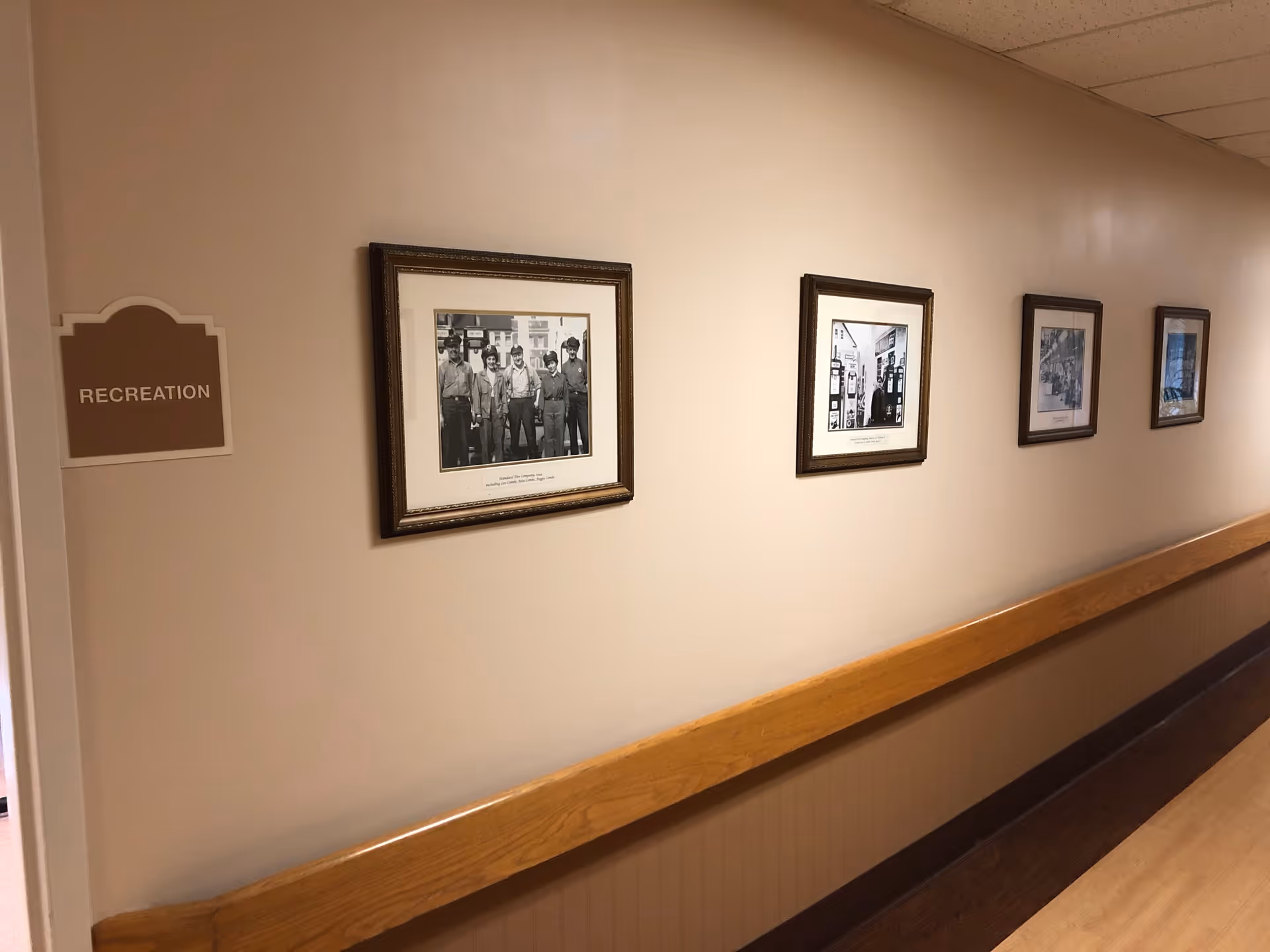 An interior hallway of a senior living facility with framed black-and-white photos along the wall and a wooden handrail.
