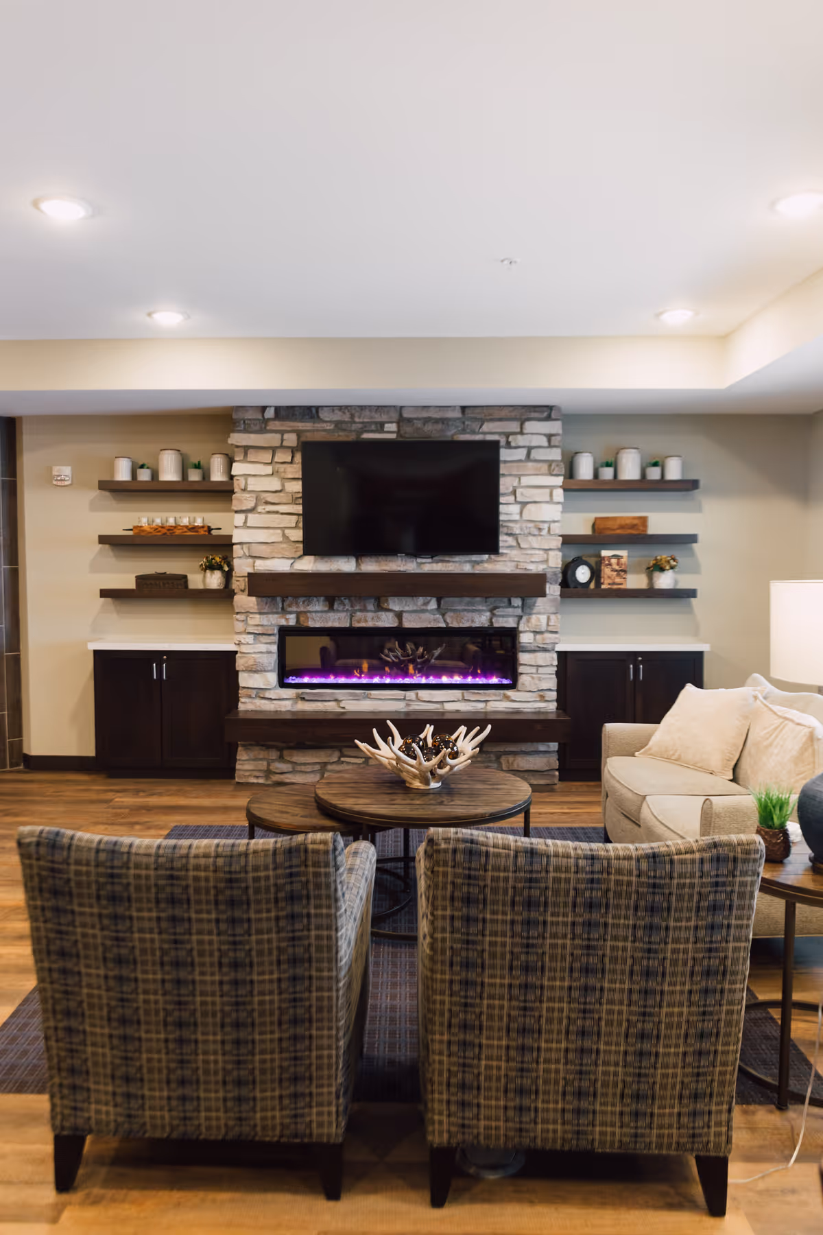 A cozy living room area featuring two plaid armchairs facing a stone fireplace with a modern electric fire and a mounted flat-screen TV above it. The fireplace wall has dark wooden shelves decorated with small plants, candles, and decorative items. To the right, there is a beige sofa with cushions and a side table with a lamp and a small plant. The room has wooden flooring and recessed ceiling lights.