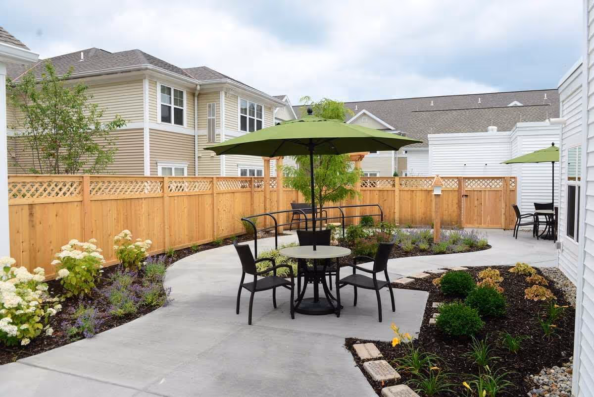 Outdoor patio area at a senior living facility with a concrete walkway, black metal table and chairs under a green umbrella, surrounded by landscaped flower beds and wooden fencing. Residential buildings are visible in the background under a cloudy sky.