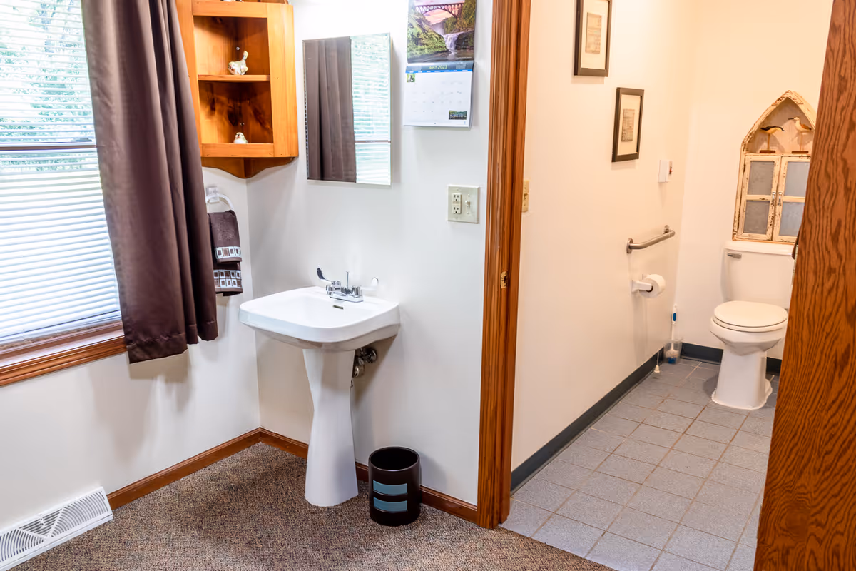 A small accessible bathroom with a pedestal sink, mirror, window with curtain, and a toilet in an adjacent tiled area.