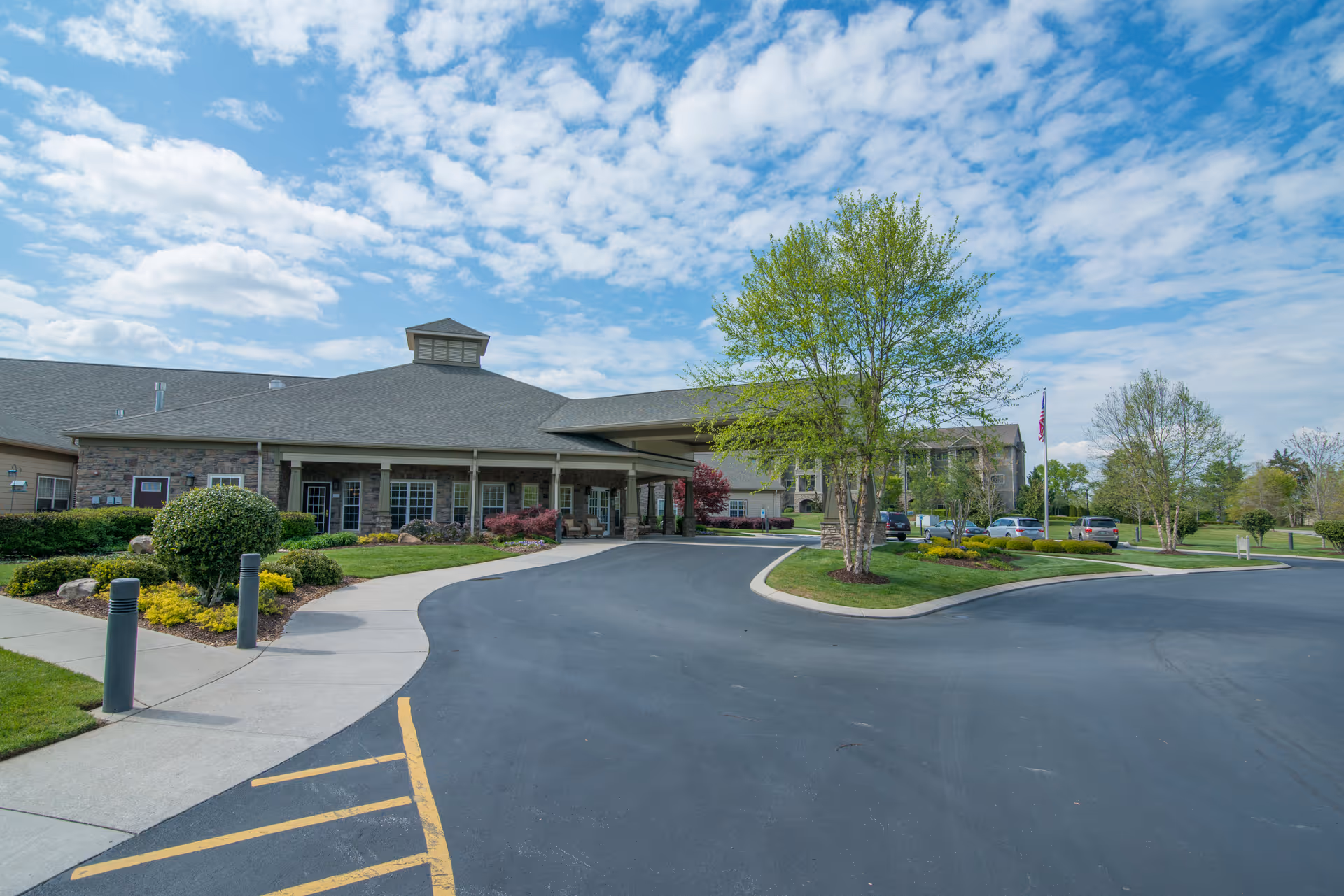 Front entrance and porte-cochère of a senior living facility with a curved driveway, landscaped grounds, and an American flag under a partly cloudy sky.