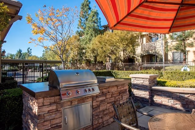 Outdoor patio with a built-in stainless steel grill set into stone counters, a round table and striped umbrella in front of a residential building.