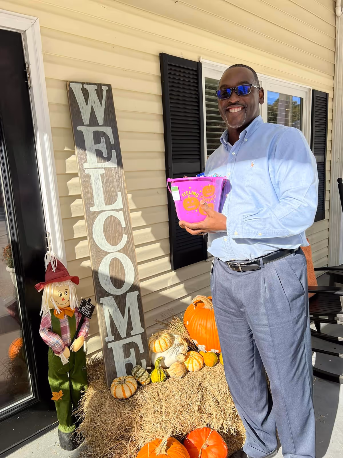 A smiling man stands on a decorated front porch holding a purple Halloween bucket next to a large "WELCOME" sign and pumpkins.