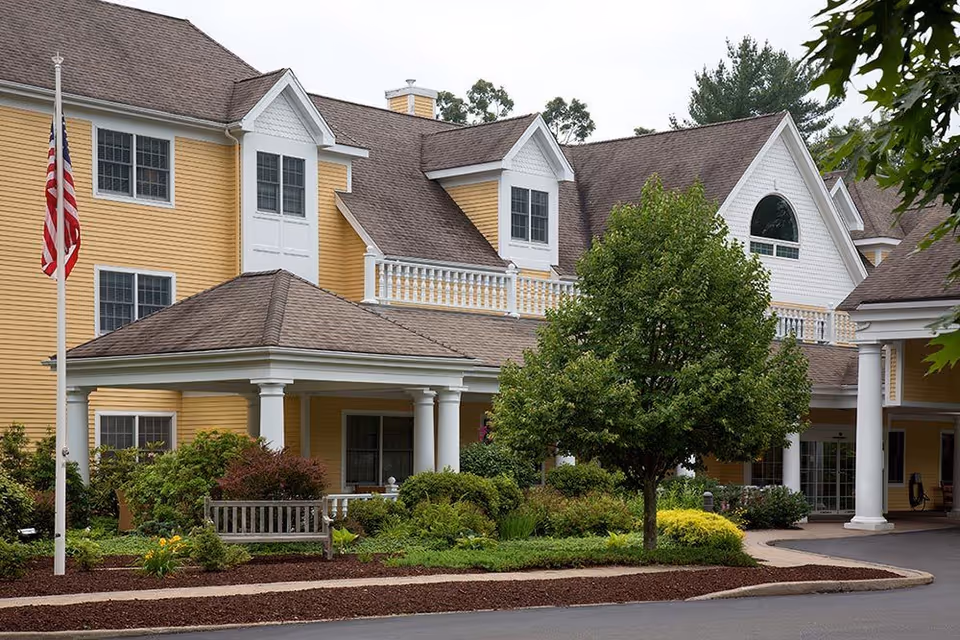 Exterior view of a senior living facility with yellow siding and white trim. The building has multiple windows, a covered entrance with white columns, a landscaped garden with bushes and a tree, and an American flag on a flagpole.