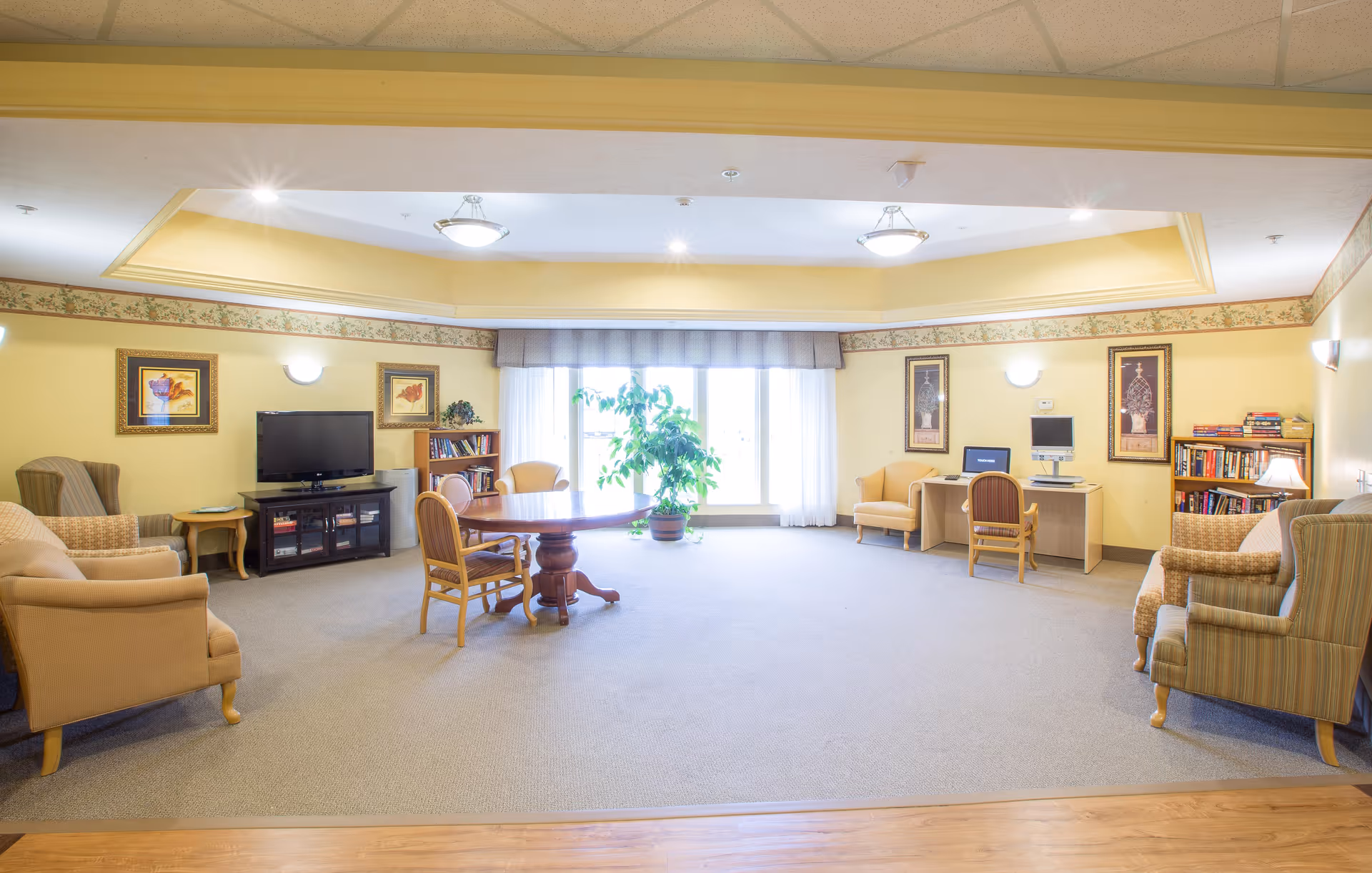 A spacious, well-lit living room area in a senior living facility with beige walls and carpeted floor. The room features several armchairs and a sofa arranged around a round wooden table. There is a TV on a stand, bookshelves filled with books, framed artwork on the walls, a computer desk with a chair, and a large window with sheer curtains letting in natural light. A potted plant is placed near the window.