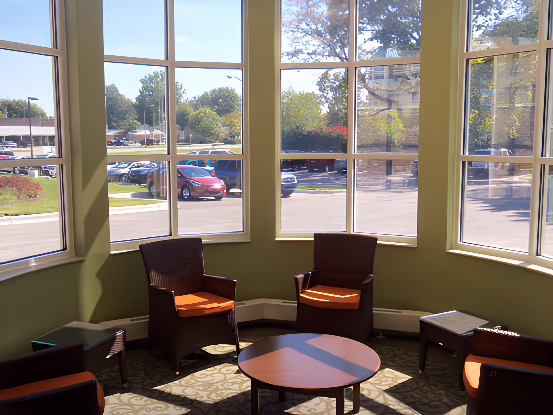 A bright seating area inside a building with large windows showing a parking lot and trees outside. The room has four wooden chairs with orange cushions arranged around a round wooden table, and two small side tables. Sunlight casts shadows on the carpeted floor.
