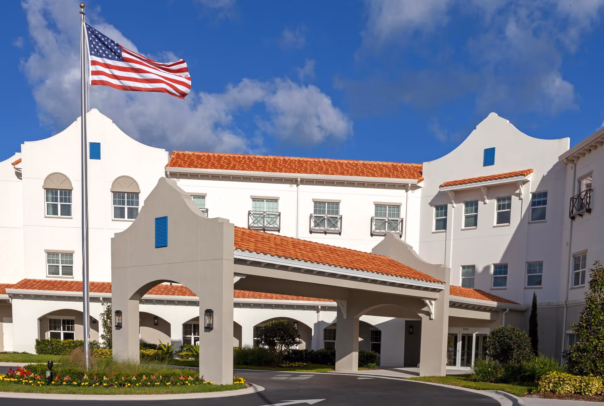 Front exterior of a white Mediterranean-style building with a covered porte-cochère and an American flag flying.