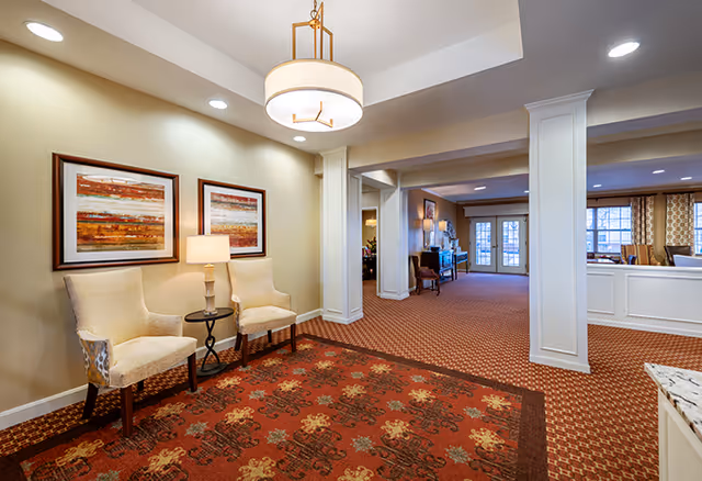 A spacious and well-lit interior common area in a senior living facility featuring two beige upholstered chairs with a small round table and lamp between them, two framed abstract paintings on the wall, a patterned carpet, white columns, and a chandelier overhead. In the background, there is additional seating and large windows allowing natural light to enter.