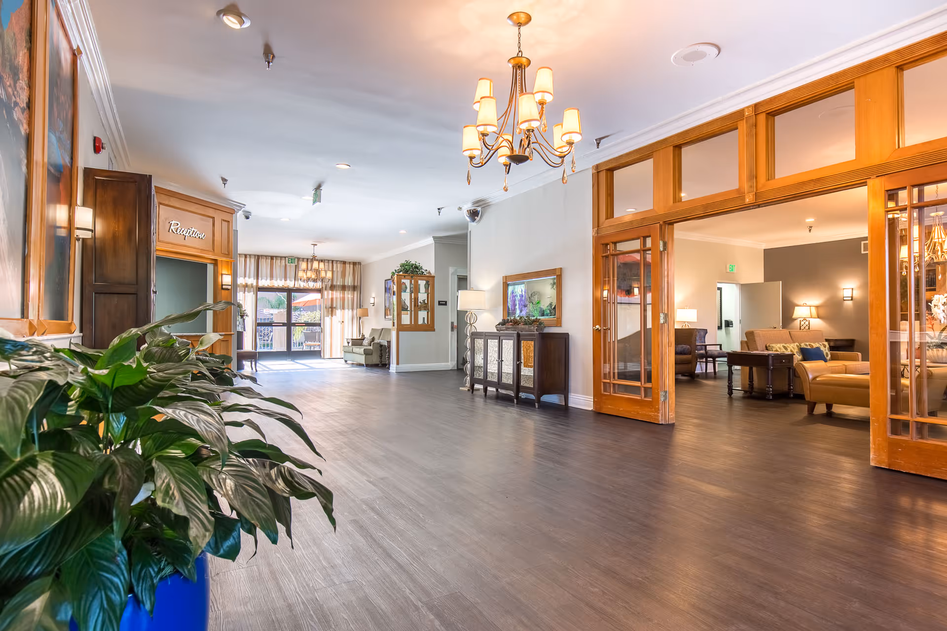Bright, spacious assisted living lobby with dark wood floors, a potted plant in the foreground, chandelier, reception area on the left, and a seating/lounge visible through wooden glass doors.