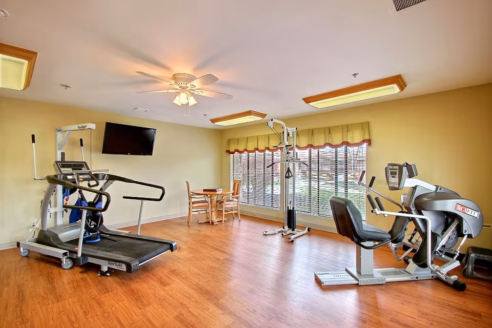 Exercise room with a treadmill, strength machine, recumbent bike, wall-mounted TV and a small table by large windows.