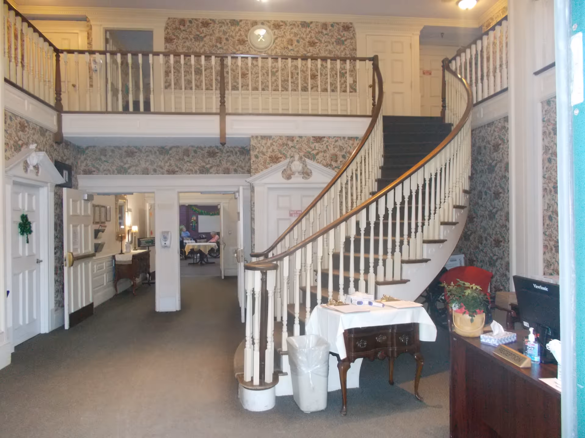 Interior view of a senior living facility lobby area featuring a curved staircase with white railings and dark wooden handrails. The walls are decorated with floral wallpaper, and there is a balcony railing above. A small table with clipboards and papers is positioned near the base of the stairs, along with a trash bin. To the right, there is a reception desk with a computer monitor, hand sanitizer, and a potted plant. The hallway beyond the staircase leads to other rooms, with some furniture and decorations visible in the distance.