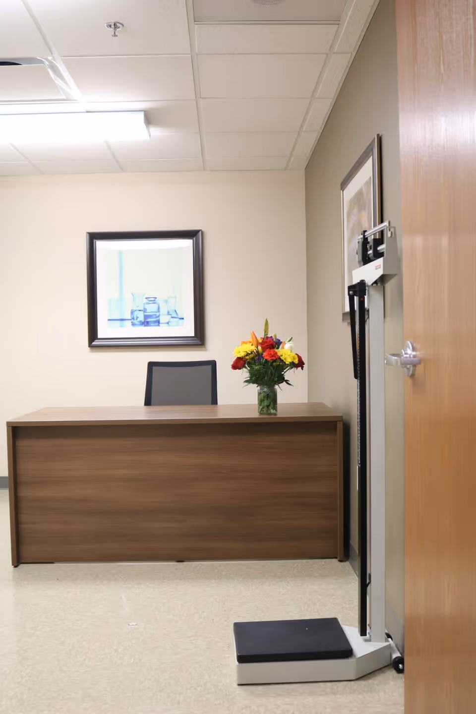A small office or consultation room with a wooden desk, a black office chair, and a vase of colorful flowers on the desk. There is a framed picture on the wall behind the desk and a medical scale with a height rod positioned near the door on the right.