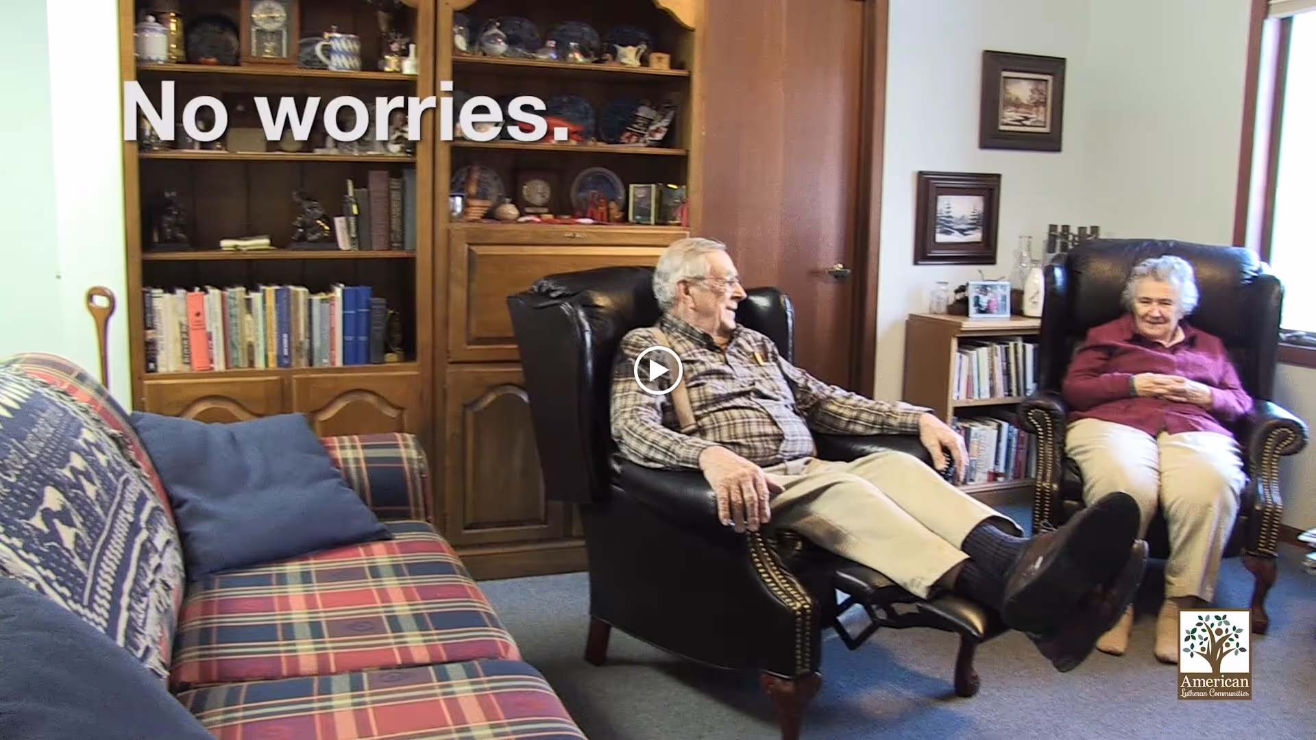 Two elderly people sitting and relaxing in a cozy living room with a plaid sofa, wooden bookshelf filled with books and decorative items, and framed pictures on the wall. The man is reclining in a black leather chair with a footrest, and the woman is seated in a similar chair nearby. Text on the image reads 'No worries.'