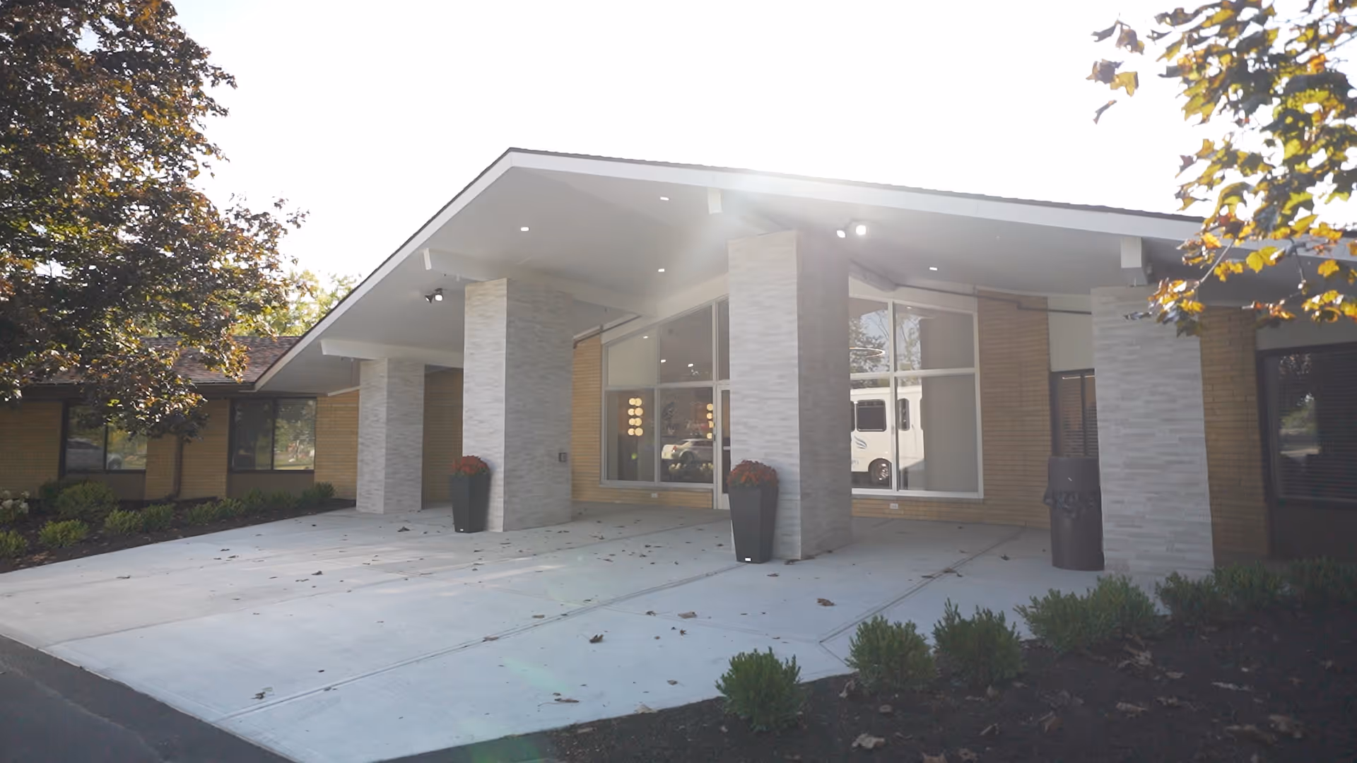 Exterior front entrance of a healthcare facility with a covered drop-off area supported by stone pillars, large windows, and landscaped bushes and trees around the building.