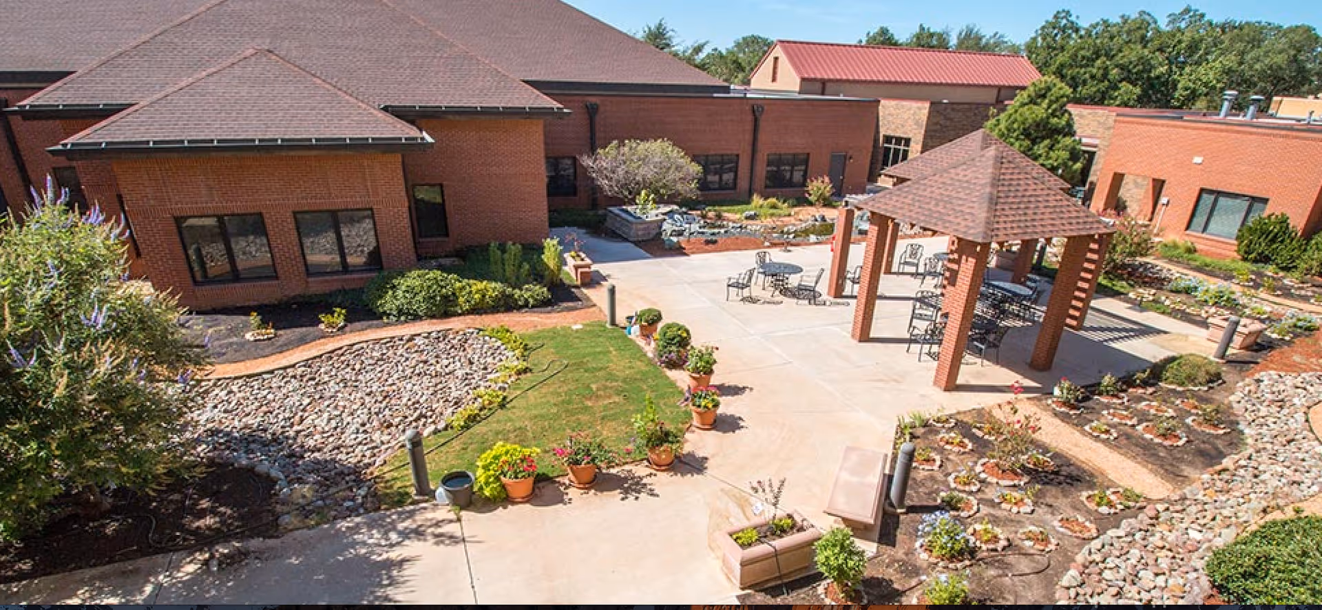 Outdoor courtyard area of a senior living facility with a gazebo, several round tables and chairs, landscaped garden beds with flowers and shrubs, potted plants along the walkway, and brick buildings surrounding the courtyard.