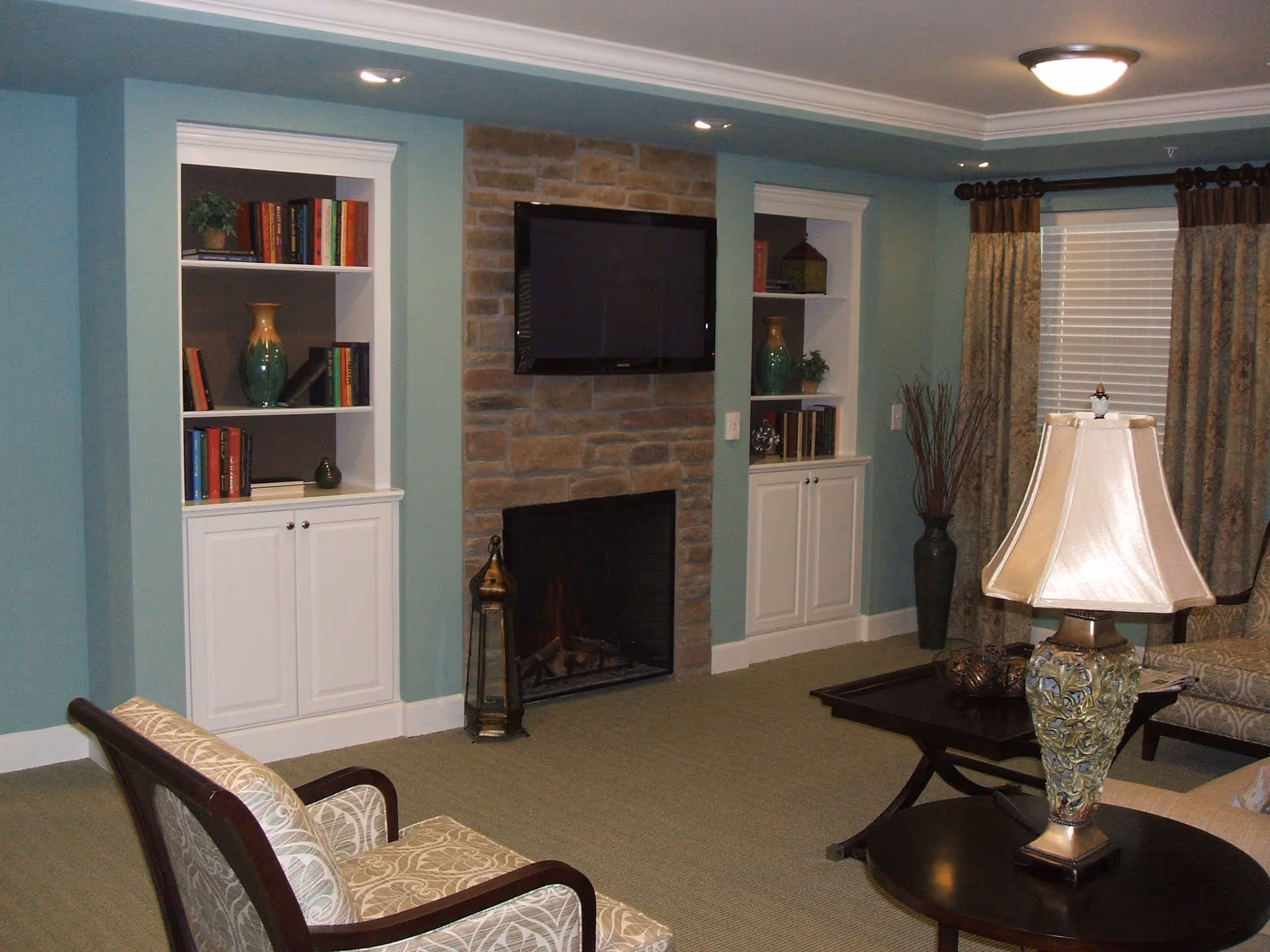 A cozy living room with light blue walls, a stone fireplace with a mounted flat-screen TV above it, built-in white shelves on either side filled with books and decorative items, a patterned armchair, a dark wooden coffee table with a decorative lamp, and a window with beige curtains.
