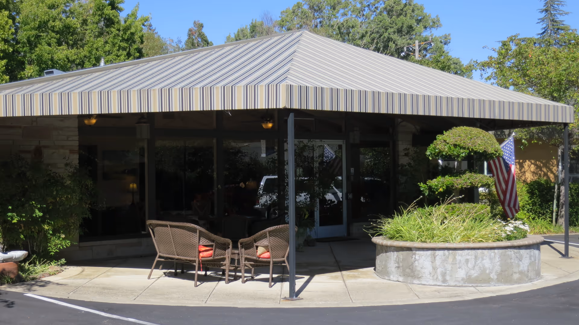Outdoor seating area at the entrance of Creekwood Care Facility with a striped canopy overhead, wicker chairs with cushions, a round concrete planter with trimmed greenery, and an American flag displayed nearby.