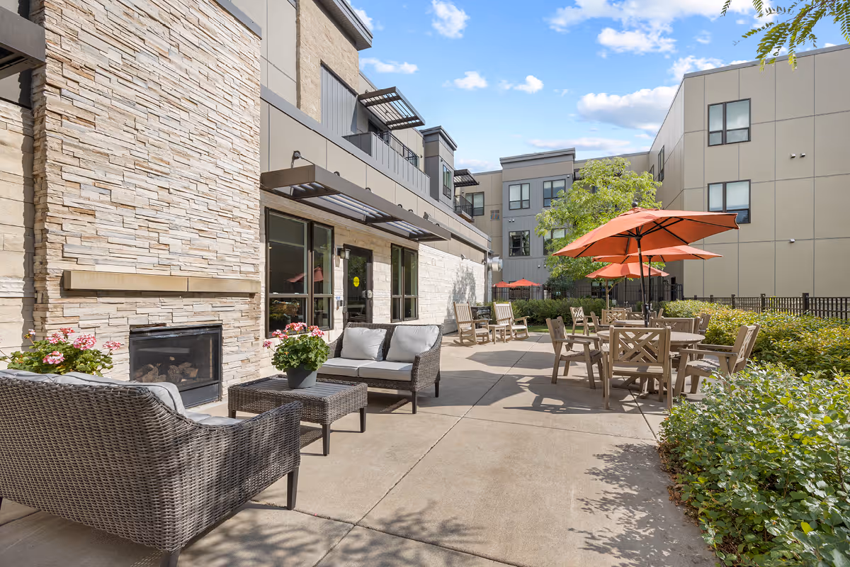 Outdoor courtyard with wicker lounge seating, a fireplace, and tables shaded by orange umbrellas beside a modern residential building.