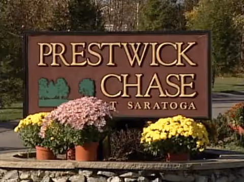 Outdoor sign reading Prestwick Chase Saratoga with yellow and pink potted flowers.