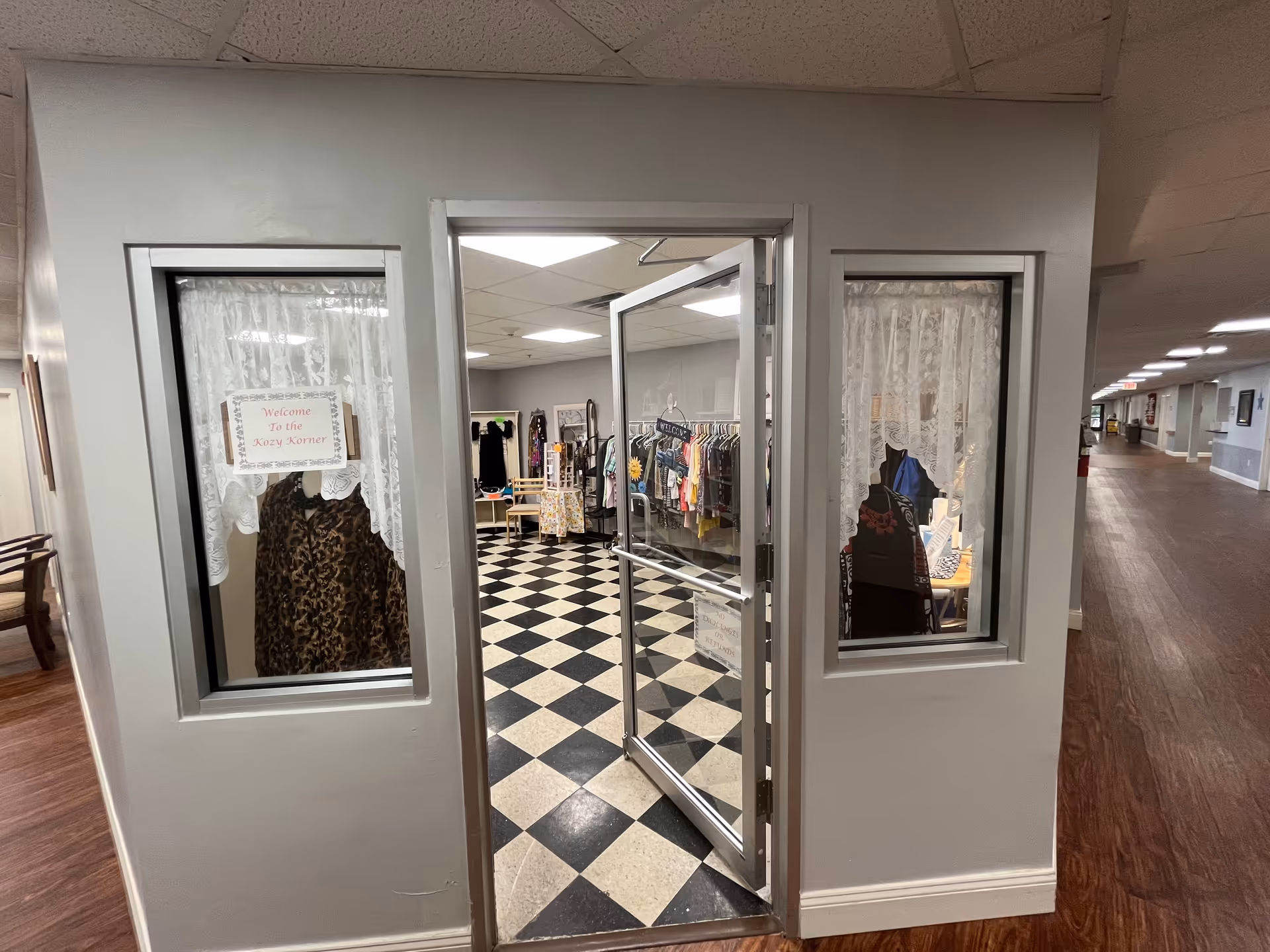 View of an entrance to a small boutique or shop area inside a senior living facility. The entrance has a glass door and two windows with white lace curtains. Inside, there are racks of clothing and a checkered black and white floor. A sign on the left window reads 'Welcome to the Kozy Korner'. The hallway outside has wood flooring and light gray walls.