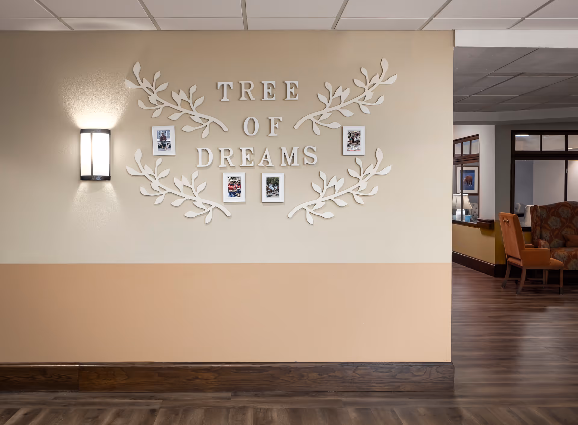 Interior wall with decorative letters spelling 'TREE OF DREAMS' surrounded by white leaf designs and small framed photos. A wall-mounted light fixture is on the left side, and a seating area with chairs and a lamp is visible in the background to the right.