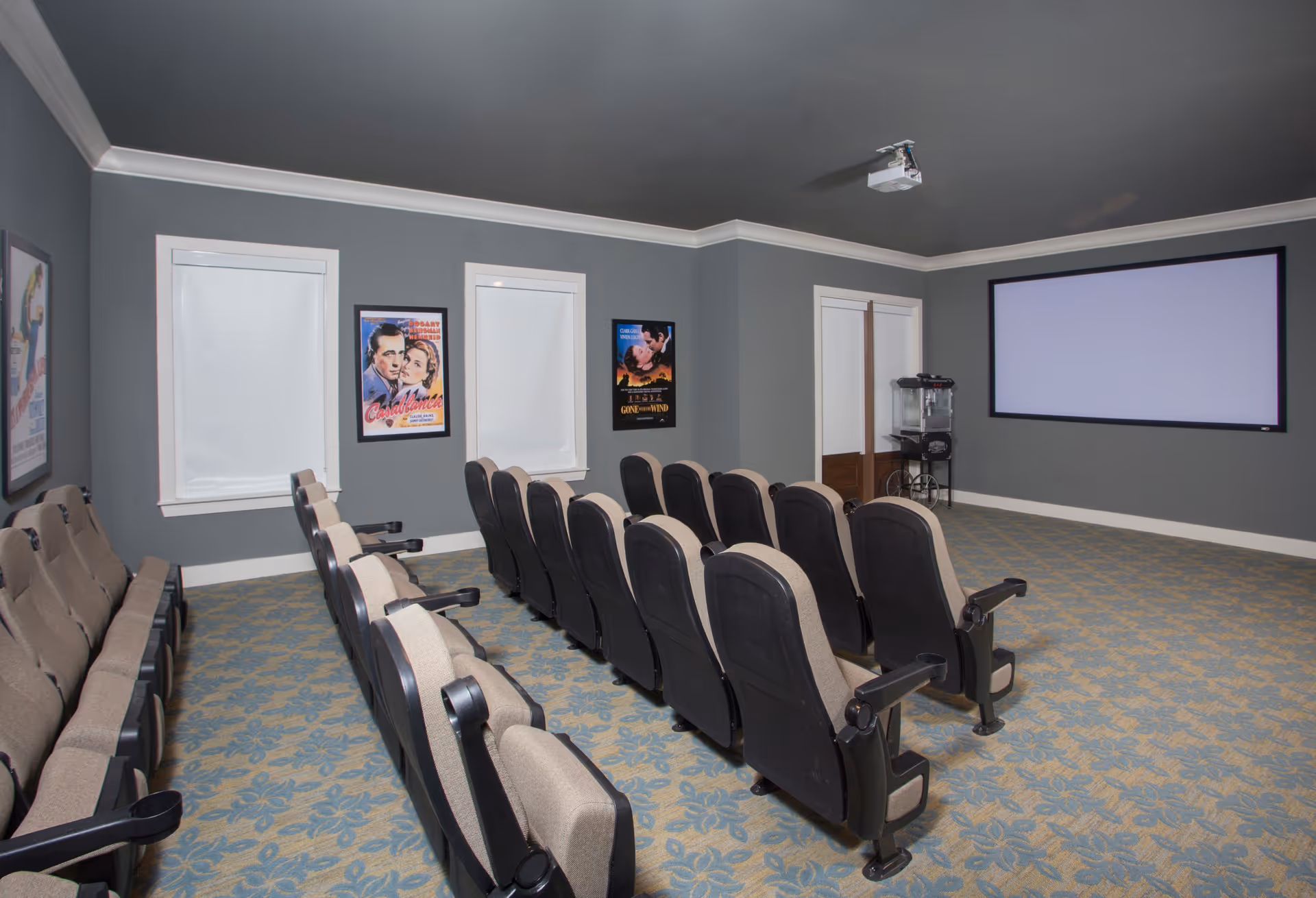 A small movie theater room with two rows of cushioned seats facing a large blank projection screen. The walls are painted gray and decorated with framed classic movie posters. There is a popcorn machine on a cart near the back wall, and a ceiling-mounted projector is visible.