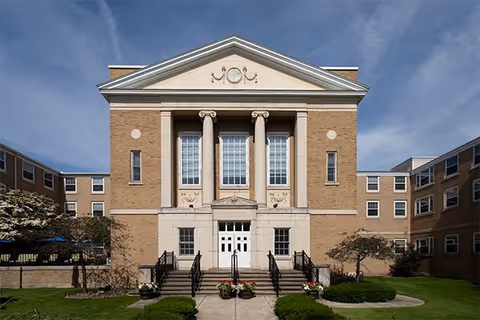 Front exterior view of a large, classical-style brick building with tall white columns, multiple windows, and a central entrance with stairs leading up to double doors. The building is surrounded by green grass, small trees, and shrubs under a partly cloudy sky.