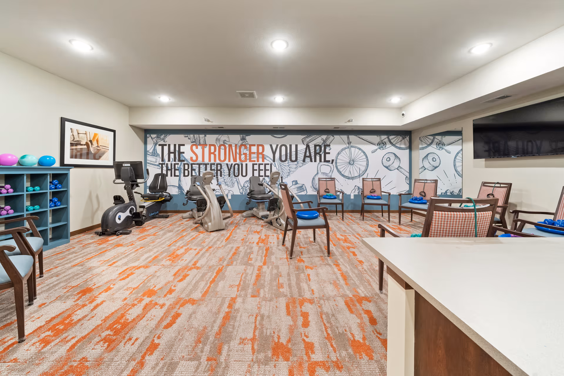 Fitness room with stationary bikes, chairs, small weights and a motivational wall mural that reads 'THE STRONGER YOU ARE, THE BETTER YOU FEEL.'
