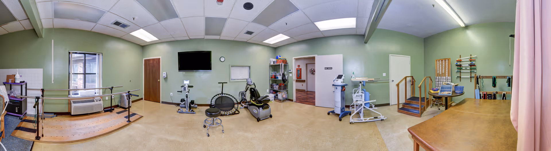 A spacious physical therapy or rehabilitation room with exercise equipment including a stationary bike, a mini trampoline, parallel bars for walking practice, a step platform with handrails, and various therapy tools on shelves and walls. The room has light green walls, a large window, ceiling lights, and a mounted TV.