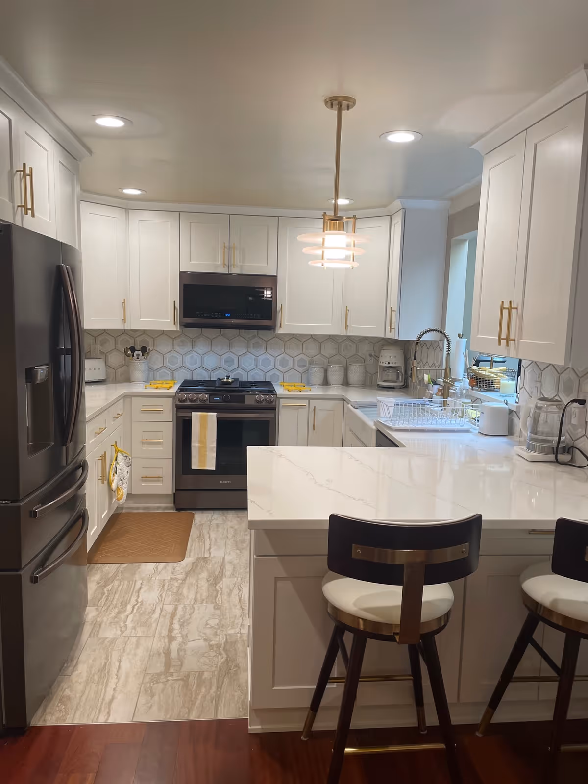 Modern kitchen with white cabinets, hexagonal tile backsplash, stainless steel refrigerator and oven, white marble countertops, two bar stools with white cushions, and a hanging light fixture above the counter.
