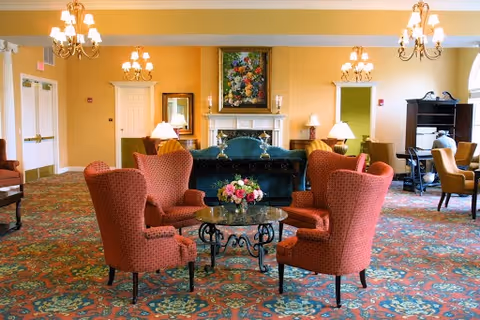 A spacious, warmly lit living room with patterned carpet, four red upholstered armchairs arranged around a glass coffee table with a floral centerpiece, a dark green sofa, a fireplace with a colorful floral painting above it, and multiple chandeliers hanging from the ceiling.