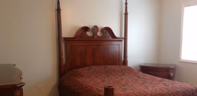 A bedroom with a large wooden four-poster bed covered with a patterned red and green bedspread. There is a matching wooden nightstand to the right of the bed and a window with white blinds letting in natural light.