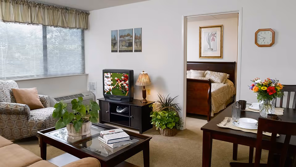 A cozy living room with a patterned armchair, a glass coffee table with a potted plant and books, a TV on a stand showing a football game, and a lamp. To the right, there is a dining table set with plates, glasses, and a vase of colorful flowers. Through an open doorway, a bedroom with a wooden bed frame and beige bedding is visible. The walls are white, and there is a window with blinds and a valance.