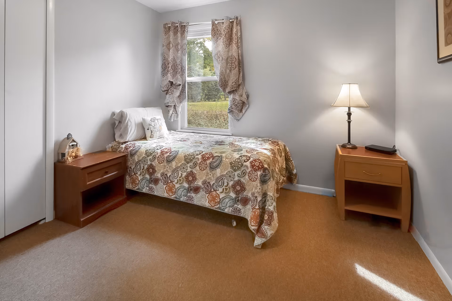 A small bedroom with a single bed covered in a colorful patterned quilt. There is a window with patterned curtains above the bed. On either side of the bed are wooden nightstands; the left one has a small decorative item and the right one has a lamp and a small black object. The walls are light gray and the floor is carpeted in a light brown color.