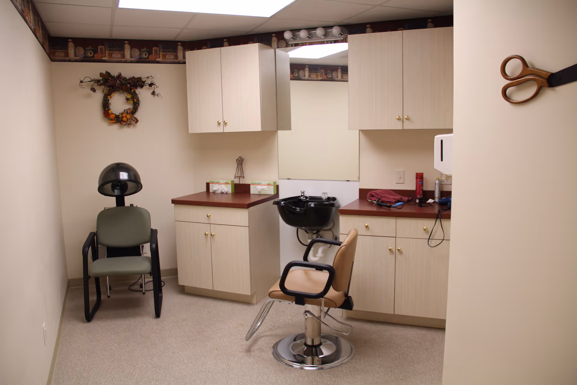 Interior of a small hair salon room with a salon chair in front of a black hair washing sink, a hair dryer chair, beige cabinets with red countertops, a large mirror, and a pair of scissors hanging on the wall.