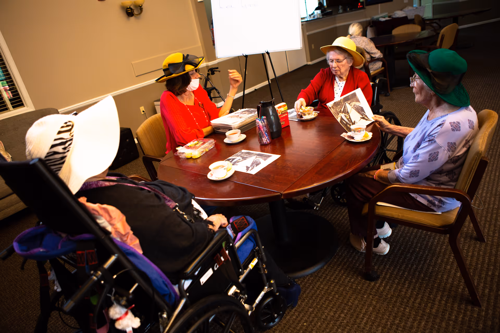 Four elderly women wearing colorful hats sitting around a wooden table in a common room. They are engaged in conversation and looking at black and white photographs. There are cups and a teapot on the table, and one woman is wearing a face mask.