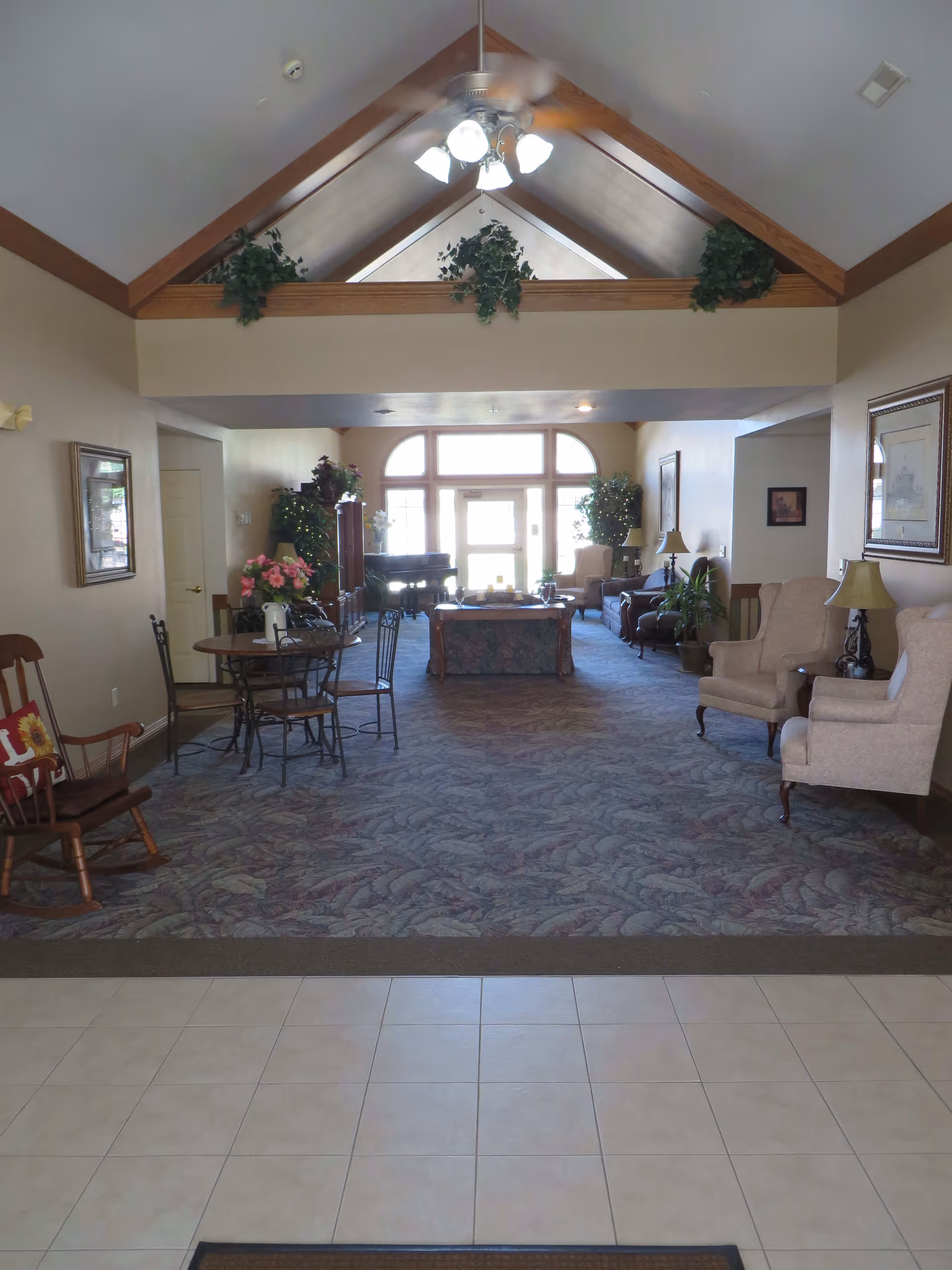 Interior view of a senior living facility common area with a vaulted ceiling and ceiling fan. The room features a carpeted floor with patterned design, several armchairs, a small round table with chairs, a rocking chair with a decorative pillow, plants, framed artwork on the walls, and large windows letting in natural light at the far end.
