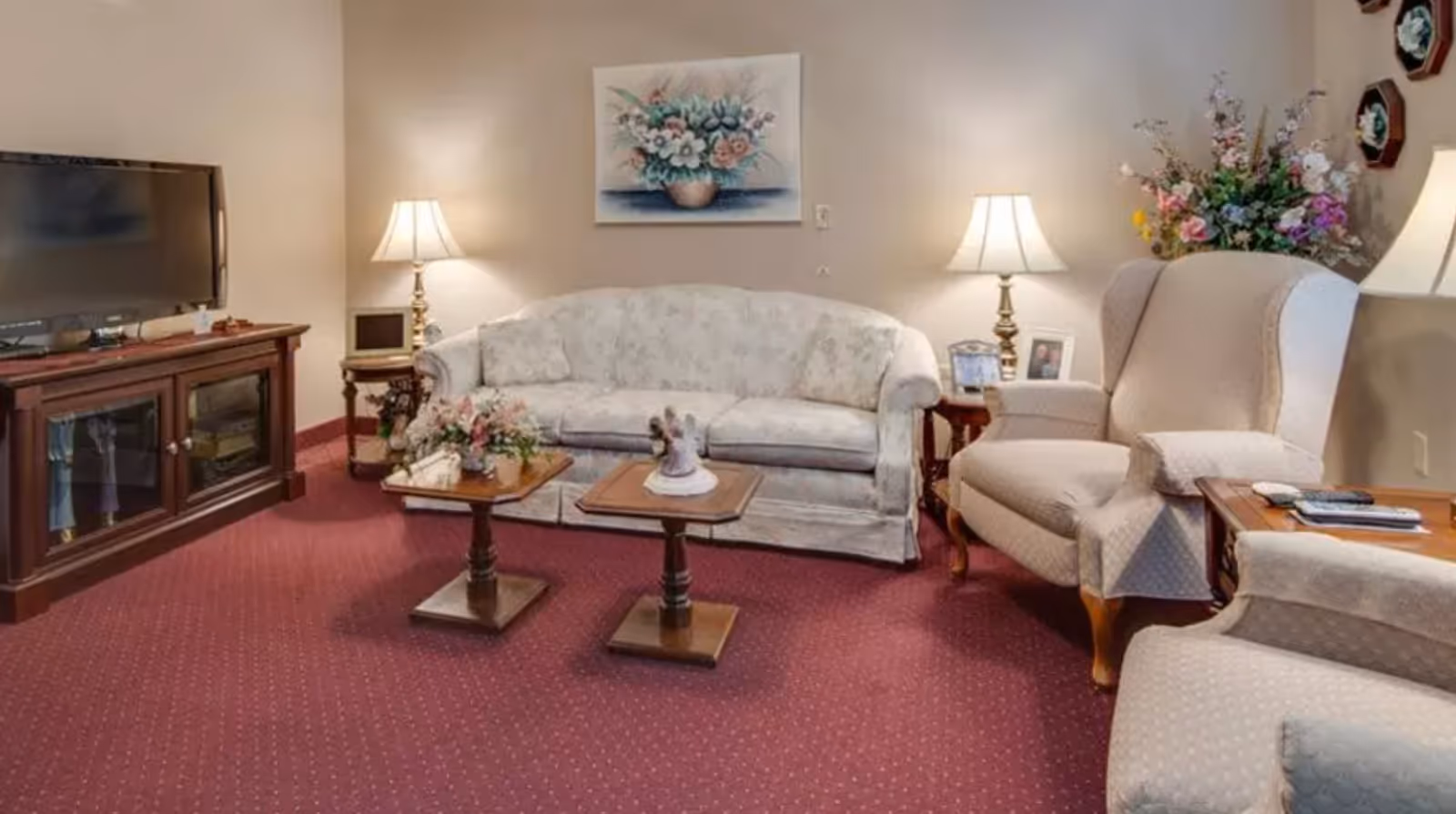 A cozy living room with a floral-patterned sofa, two beige armchairs, two wooden side tables with floral decorations, a wooden TV stand with a flat-screen TV, two table lamps on side tables, a floral painting on the wall, and a large floral arrangement in the corner. The room has a red carpet and beige walls.