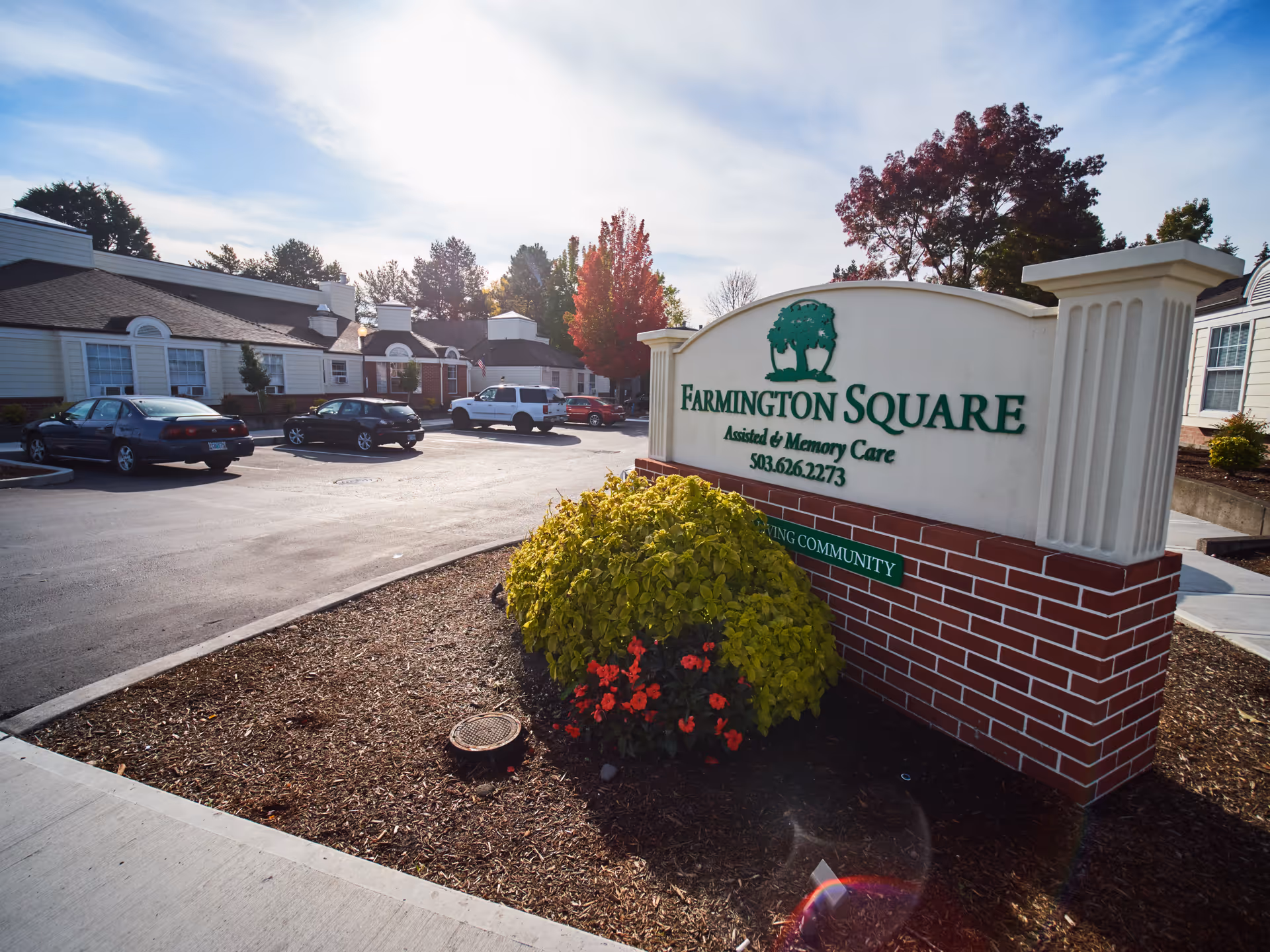 Outdoor view of Farmington Square Beaverton assisted and memory care facility sign with a landscaped area featuring green and red plants in front. Several parked cars and single-story buildings are visible in the background under a partly cloudy sky.