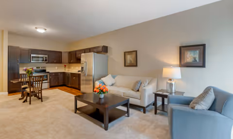 Open-plan living area with a beige sofa, armchair and coffee table adjacent to a kitchen with dark cabinets and stainless steel appliances.