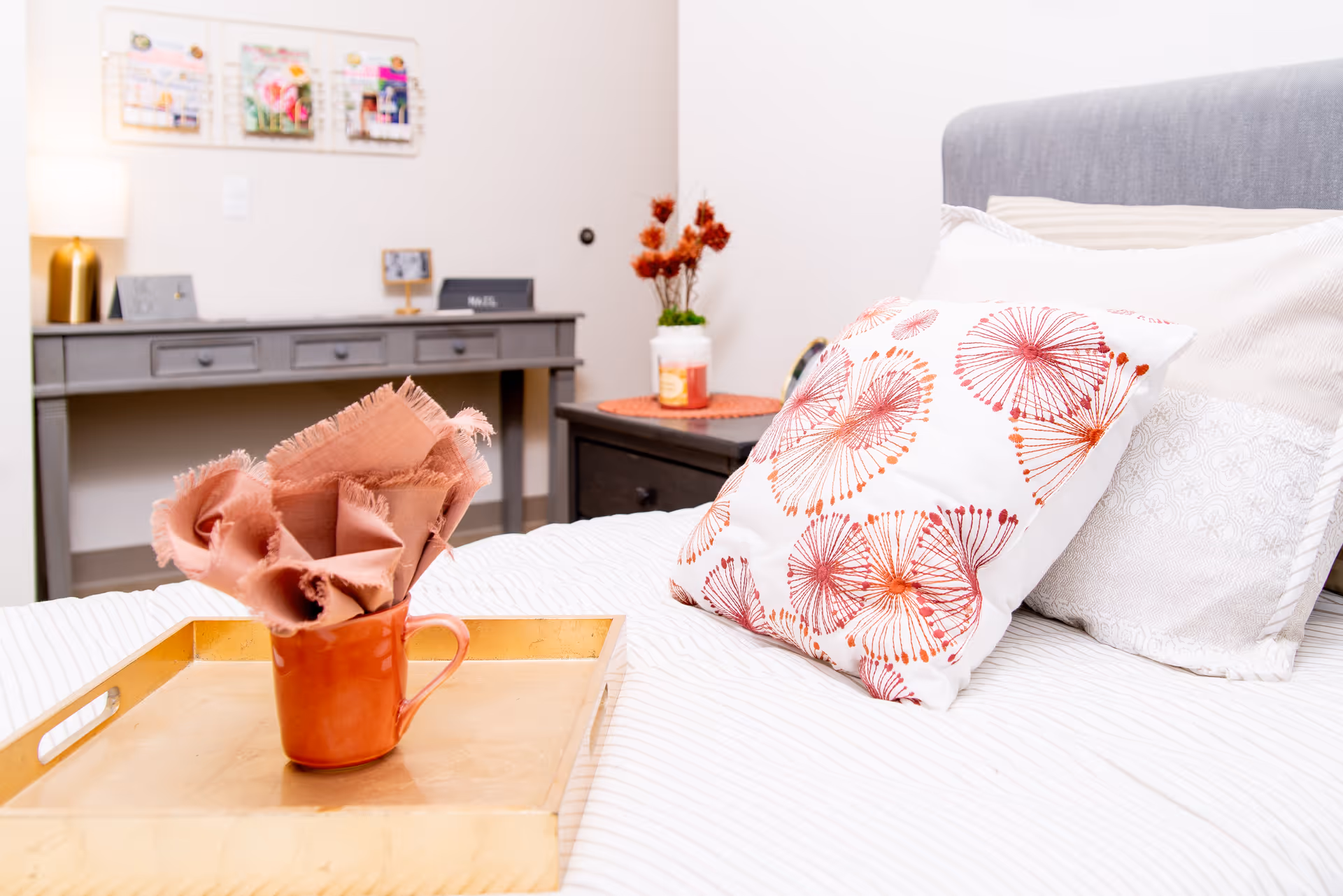 A cozy bedroom scene featuring a bed with white and patterned pillows, a wooden tray holding a red mug with a folded pink cloth, a gray desk with drawers in the background, a lamp, and decorative items including a small plant and framed pictures on the wall.