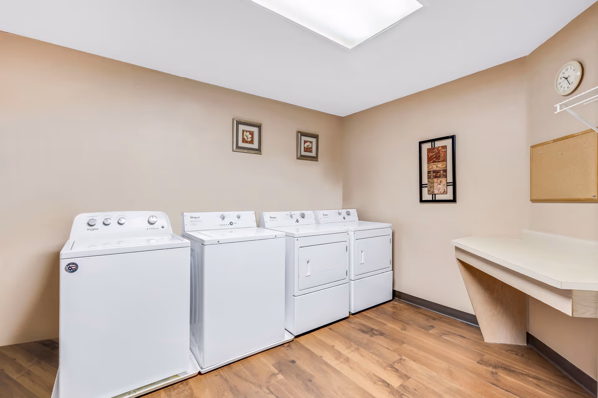 A laundry room with four white washing machines and dryers lined up against a beige wall. The room has wooden flooring, a white countertop on the right side, a clock, a corkboard, and framed artwork on the walls.