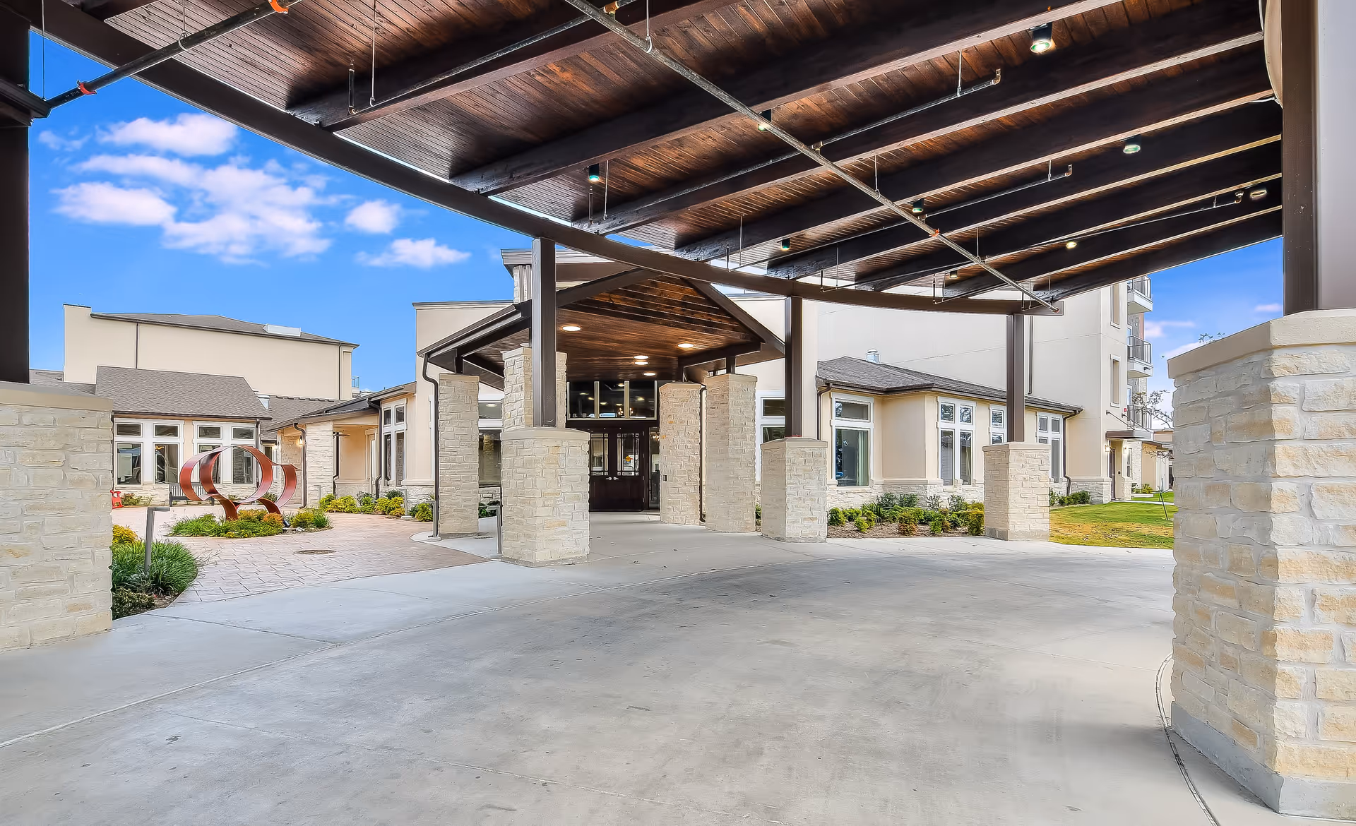 Covered entrance area of a senior living facility with stone pillars supporting a wooden roof structure, leading to the main building entrance. The building has beige walls and multiple windows, with a small landscaped garden area visible to the left featuring a modern metal sculpture. The sky is clear with a few clouds.